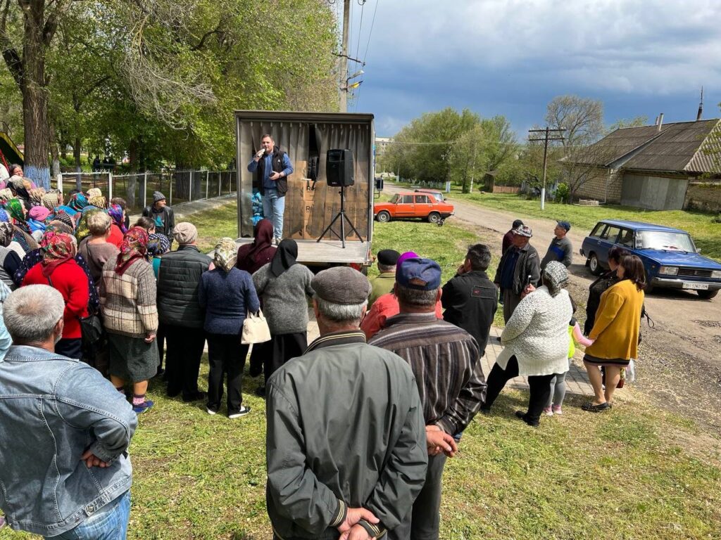 Man speaking to a crowd of Ukrainians near a dirt road. 