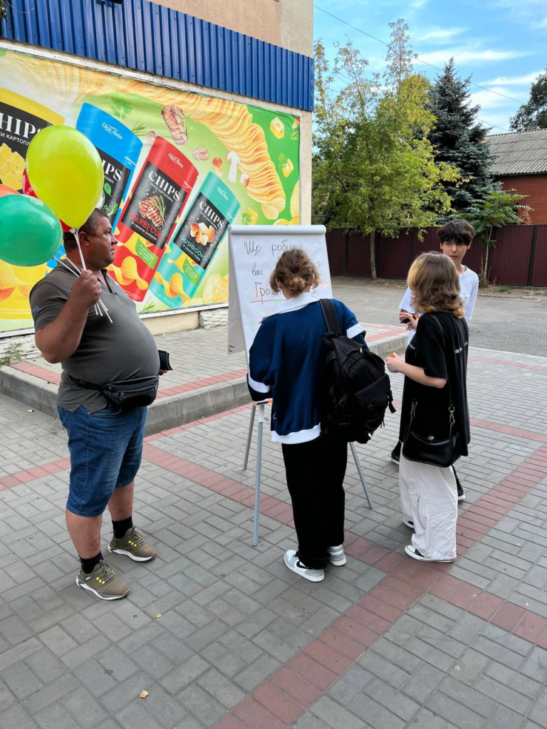 A few youth looking at a paper drawing board with an adult man nearby holding balloons.