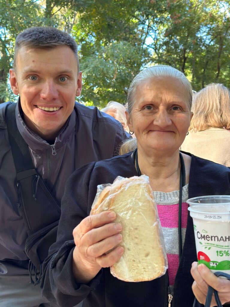 elderly woman holding a cup of warm soup and a sandwich. She is smiling. There is a man standing next to her