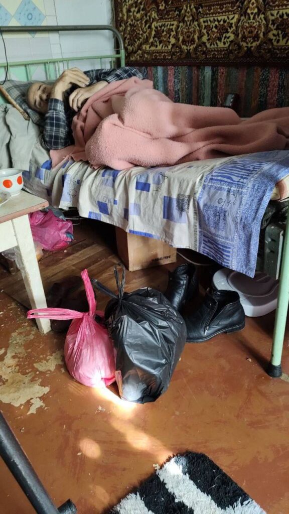 An elderly man in bed with two bags of items next to him on the floor