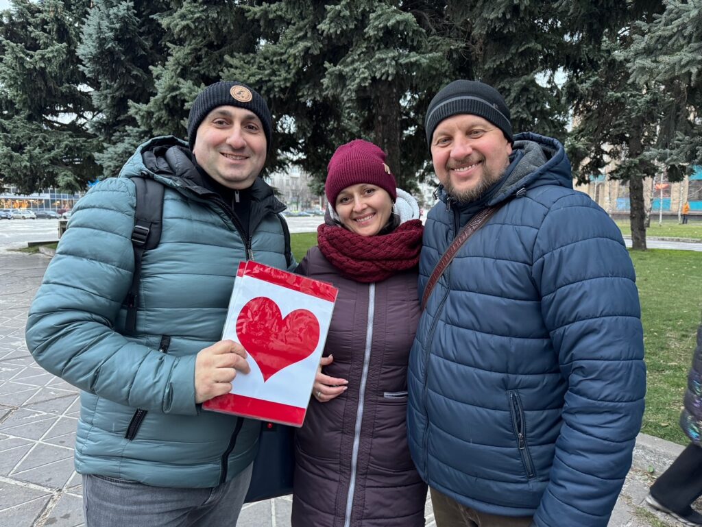 Three adults smiling at camera with red heart image in hand