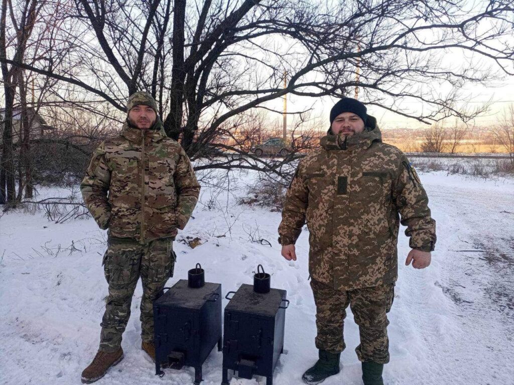 Two men in camouflage standing next to two stoves. 