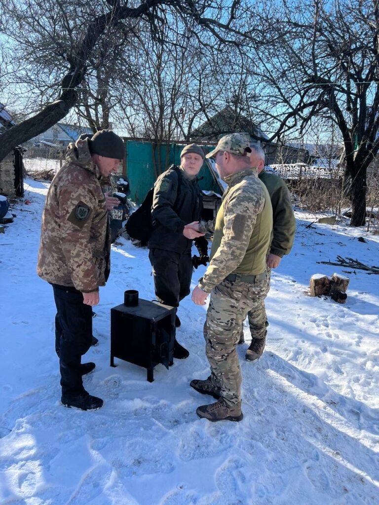 Two men in camouflage shaking hands with other men while gathered around stove. 